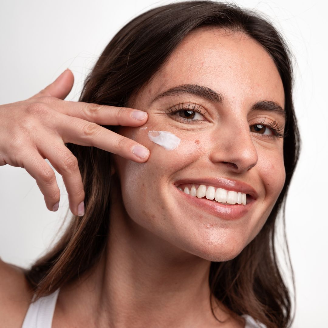 Donna applica crema sul viso. Crema bianca sulla pelle. Sorride.