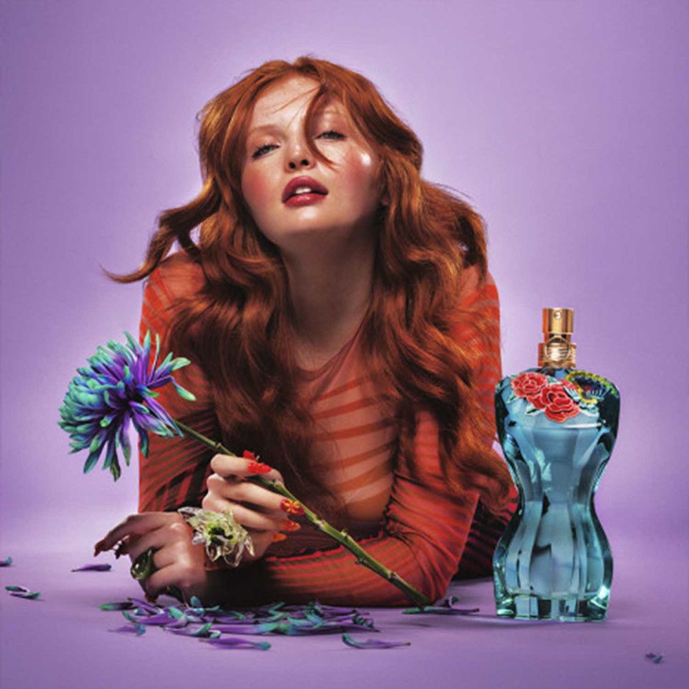 Donna con capelli rossi e top rosso. Accanto, un flacone di profumo blu a forma di corpo femminile, decorato con fiori.