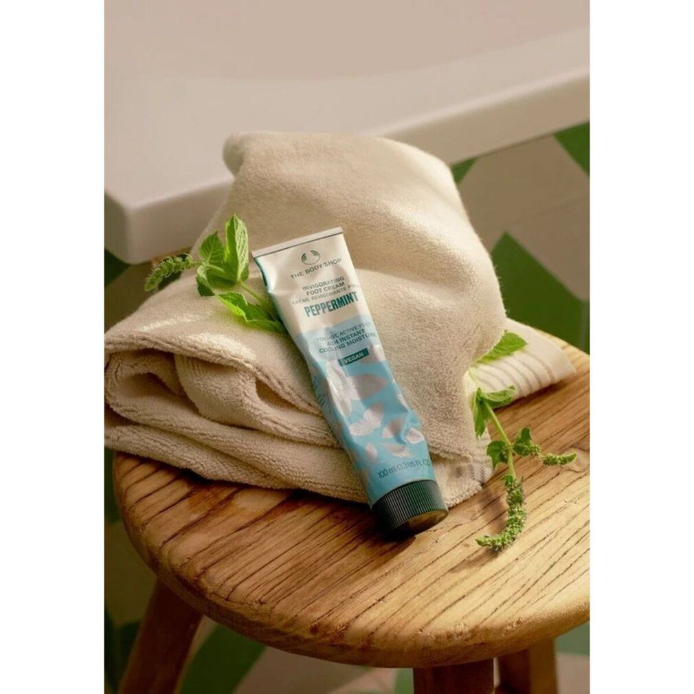 Tubo di scrub per piedi su uno sgabello con asciugamano e rametti di menta. Nome e logo del prodotto visibili.