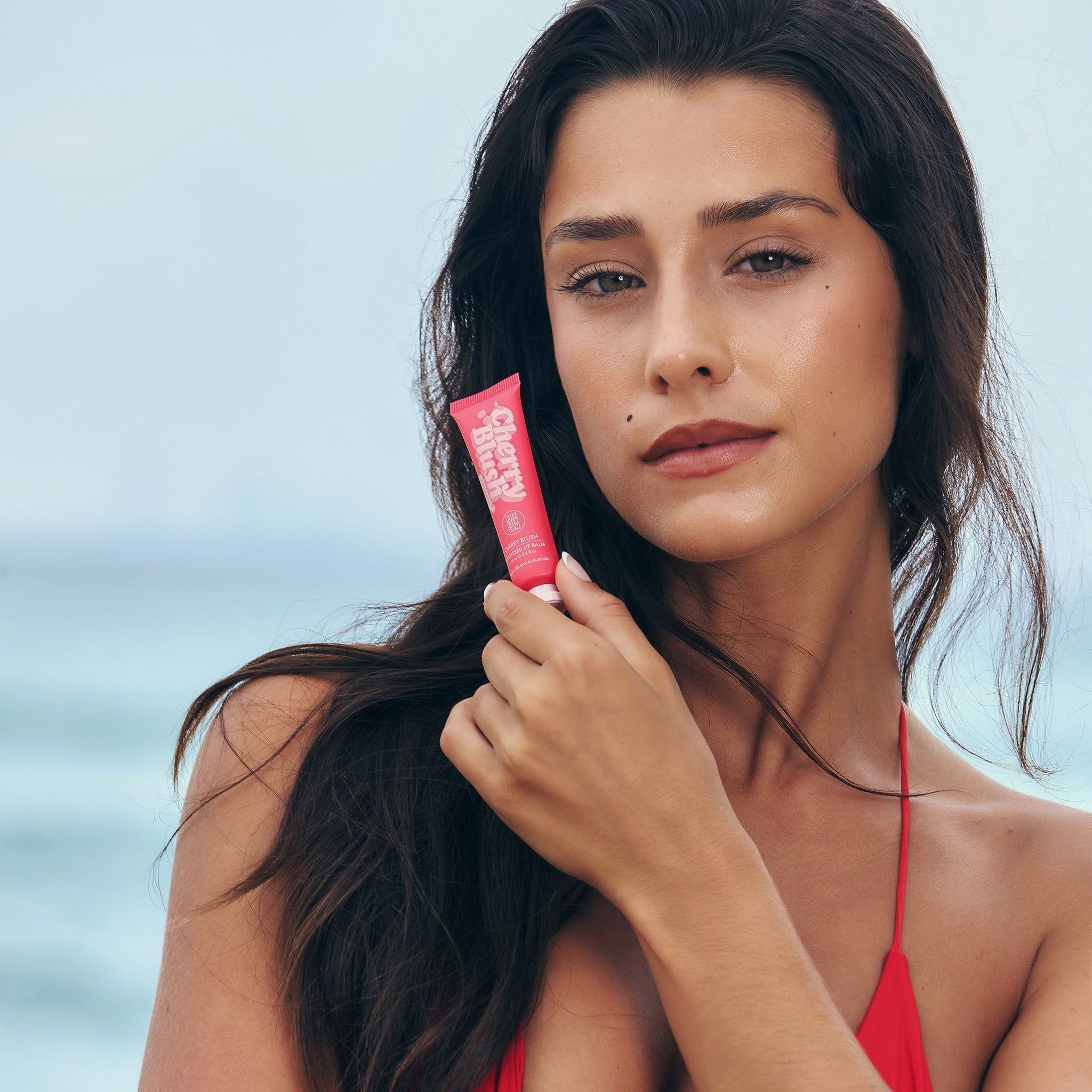Donna tiene tubo 'Cherry Blush'. Nome del prodotto e marchio visibili. Top rosso. Sfondo spiaggia.