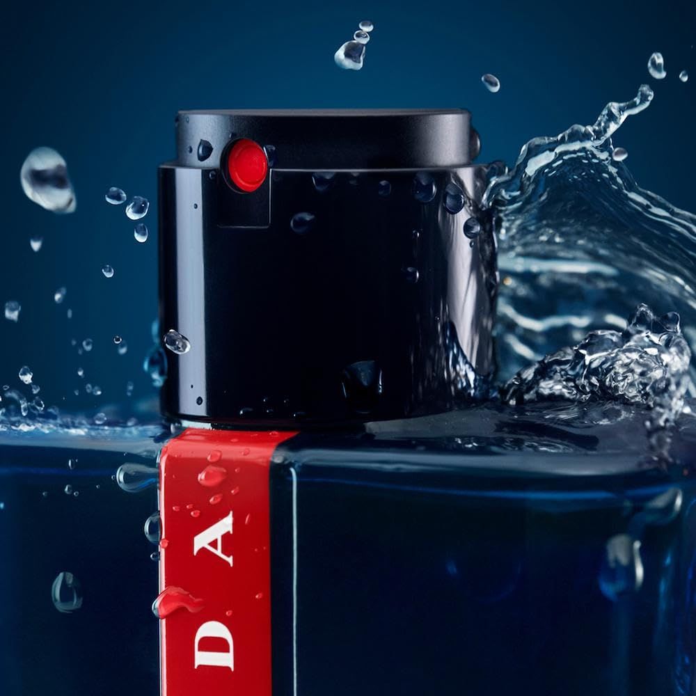 Flacone blu scuro, striscia rossa Prada. Erogatore nero. Spruzzi d'acqua. Primo piano.