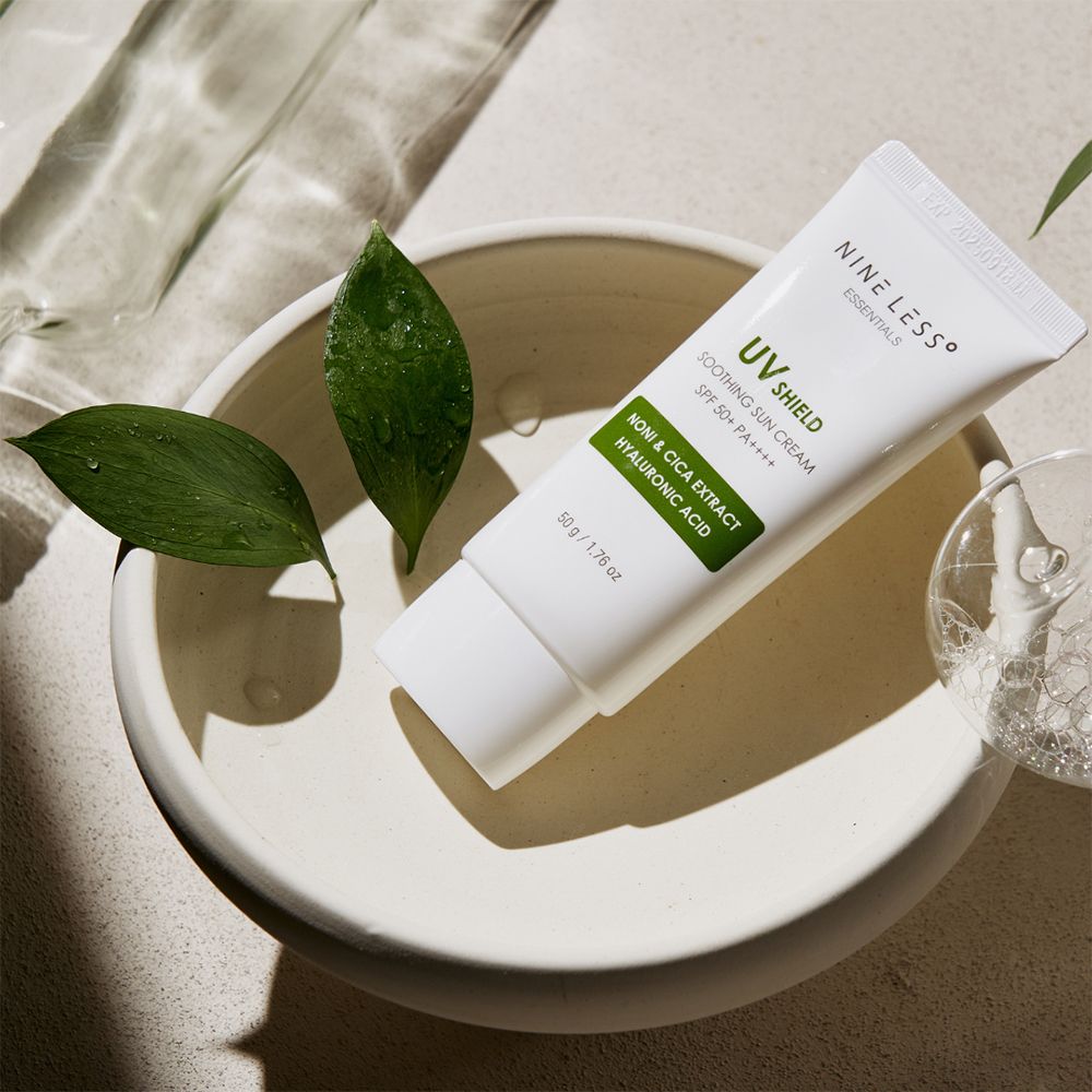 Tubo bianco in una ciotola con foglie. Scritta: UV Shield, SPF 50+ PA++++, Noni & Cica Extract, Acido Ialuronico. Marchio: NINE LESS.