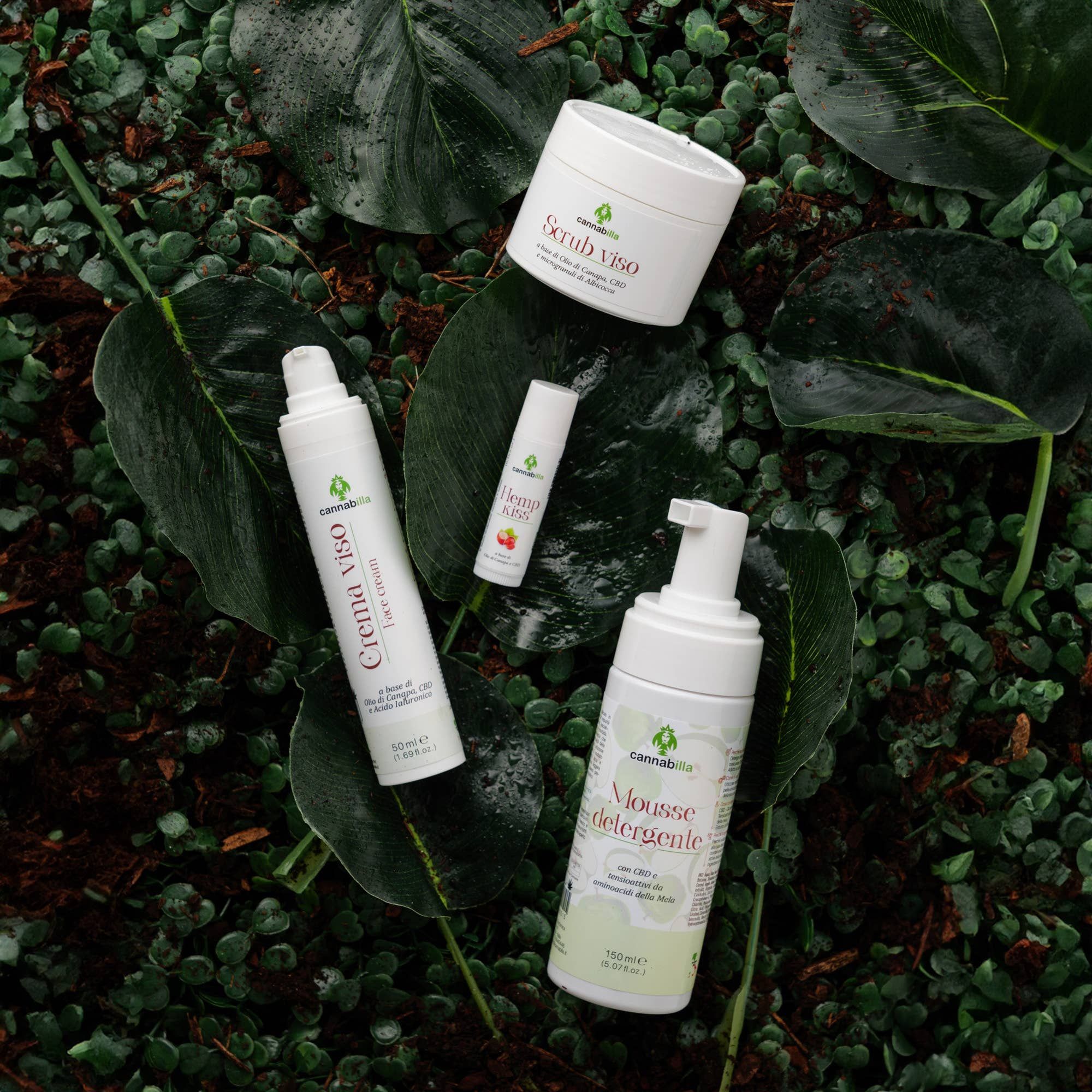 Vari prodotti Cannabilla su foglie. Include: crema viso, mousse detergente, scrub e balsamo labbra. Logo e scritta Cannabilla.