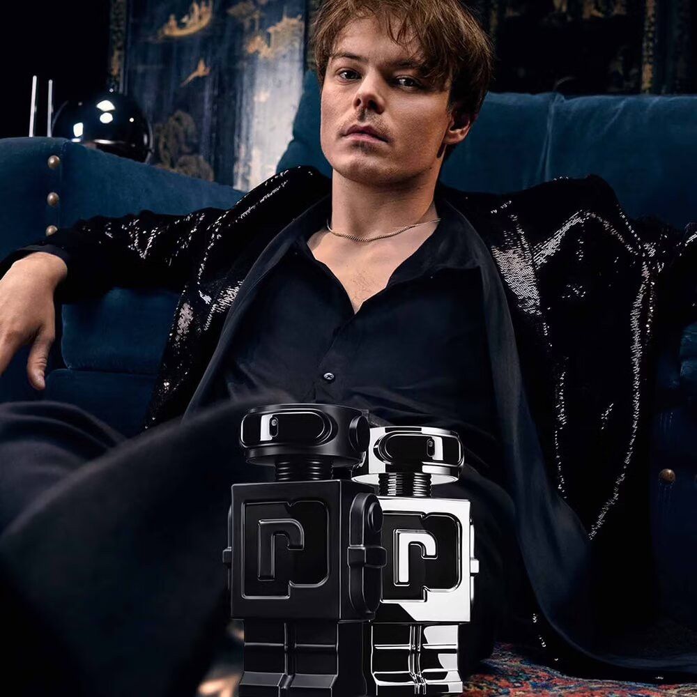 Uomo seduto su un divano, tiene due flaconi robot. Uno argentato, uno nero. Abiti neri.