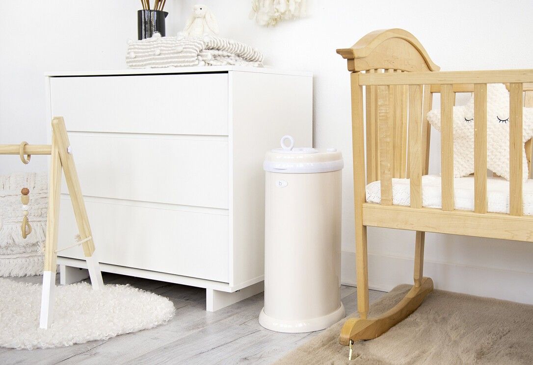 Bidone per pannolini beige. Forma cilindrica. Coperchio bianco con maniglia. Vicino a cassettiera e culla.