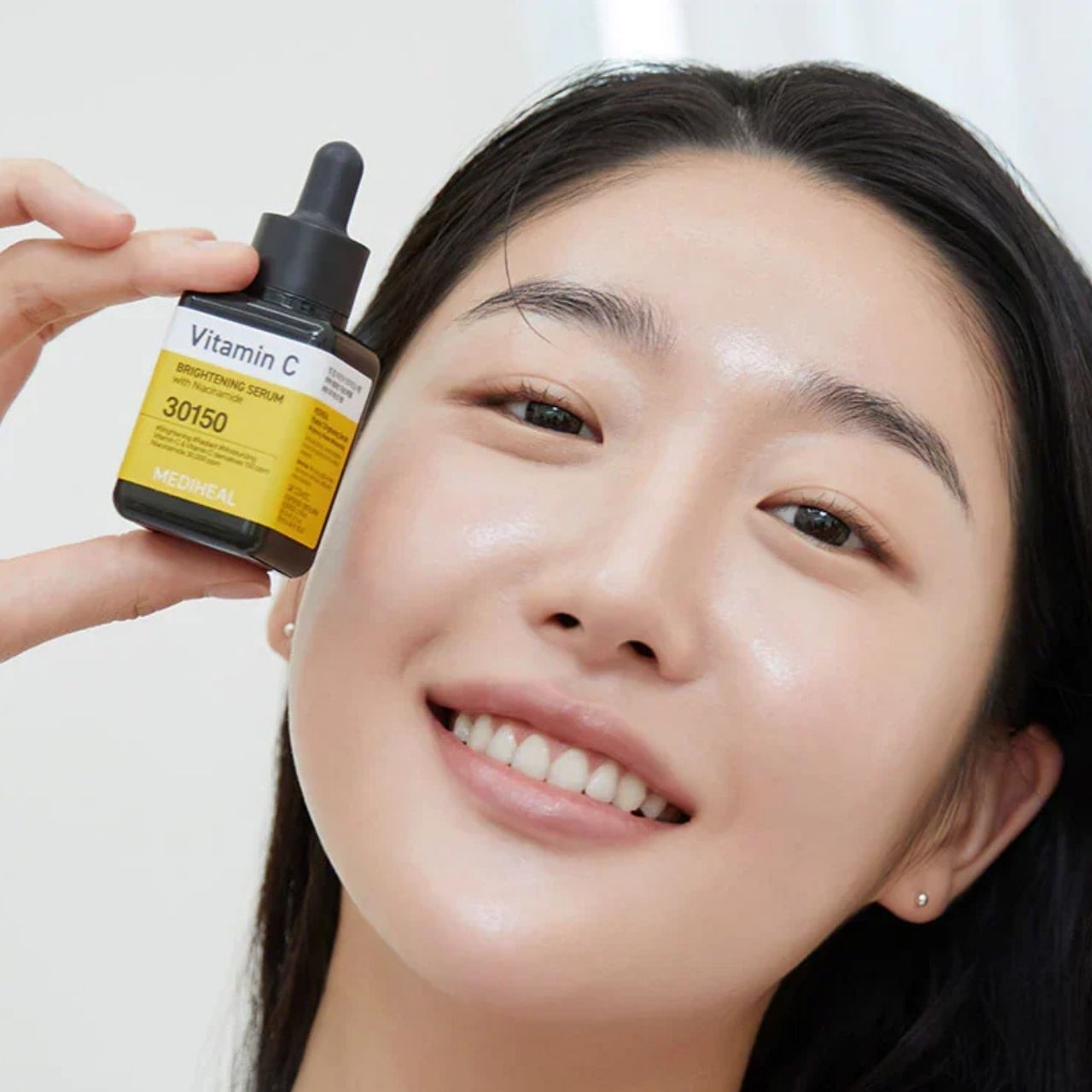 Una donna tiene un flacone di siero Mediheal Vitamina C davanti al viso. Etichetta gialla con il nome del prodotto.