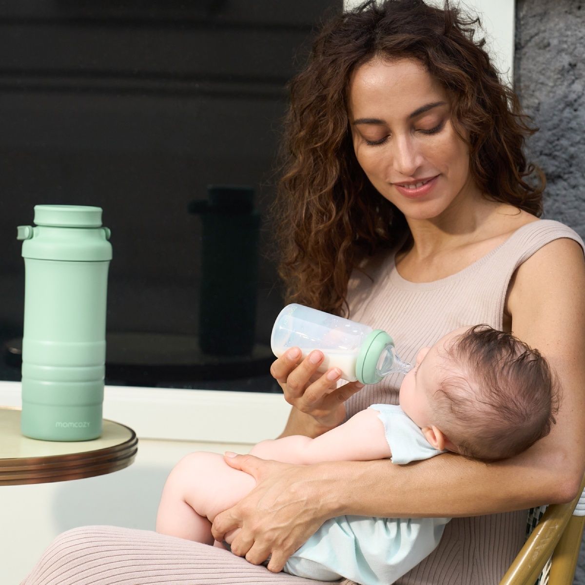 Donna che allatta il bambino con il biberon. Accanto a lei, un refrigeratore per latte materno Momcozy verde.