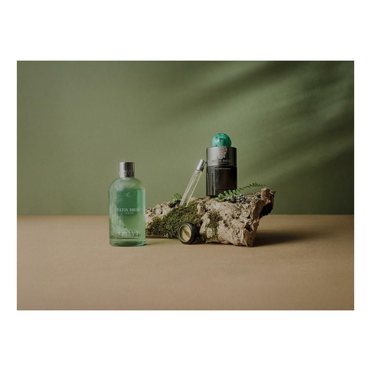 Flacone di profumo e vaporizzatore su un pezzo di legno, circondati da muschio. Sfondo verde.