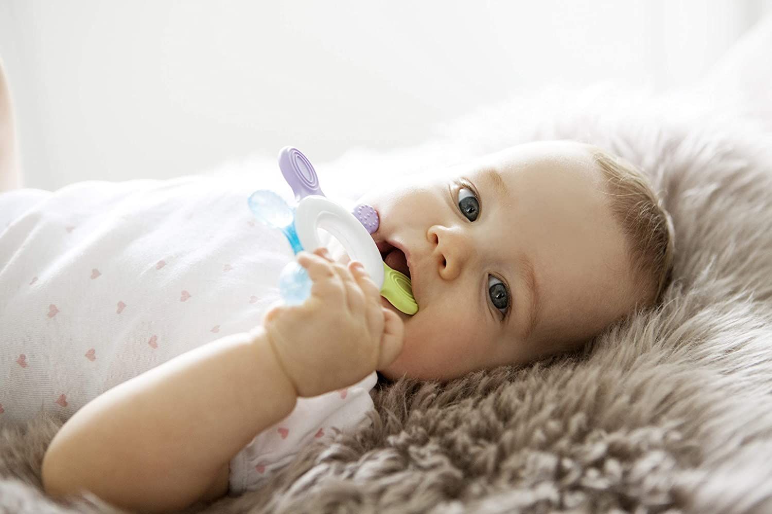 Bambino sdraiato su pelliccia, morde anello refrigerante. Anello blu, verde, viola e bianco.