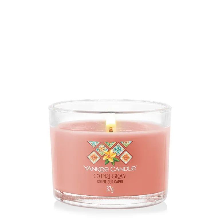 Candela rosa in contenitore di vetro. Scritta: Capri Glow, Soleil sur Capri, 37g. Uno stoppino acceso.