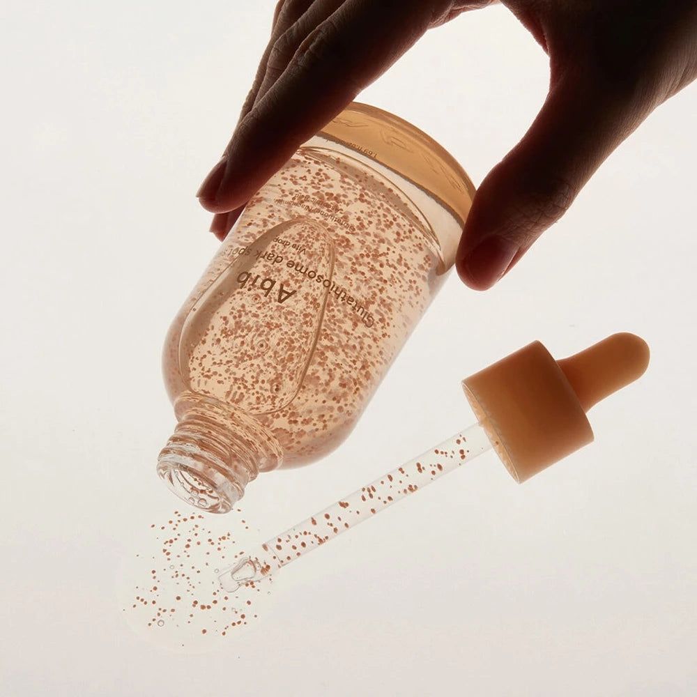 Flacone in vetro con siero, perle rosa, pipetta. Una mano tiene il flacone. Il siero viene versato su una superficie.
