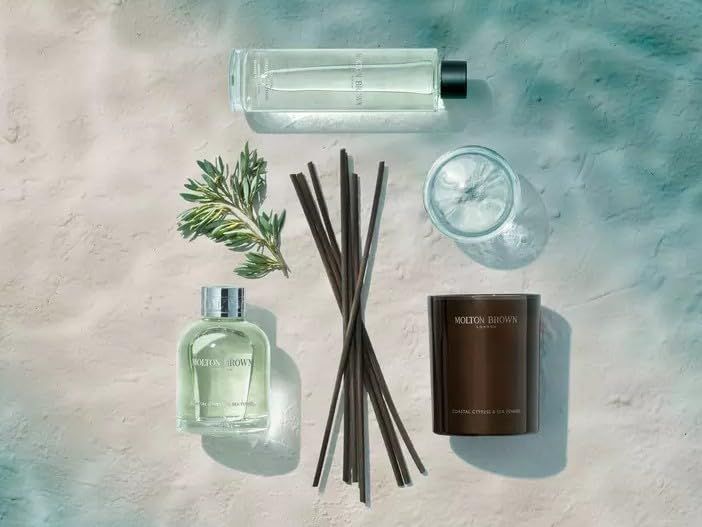 Vari prodotti Molton Brown: flacone, diffusore, candela, ramo. Su sfondo sabbia.