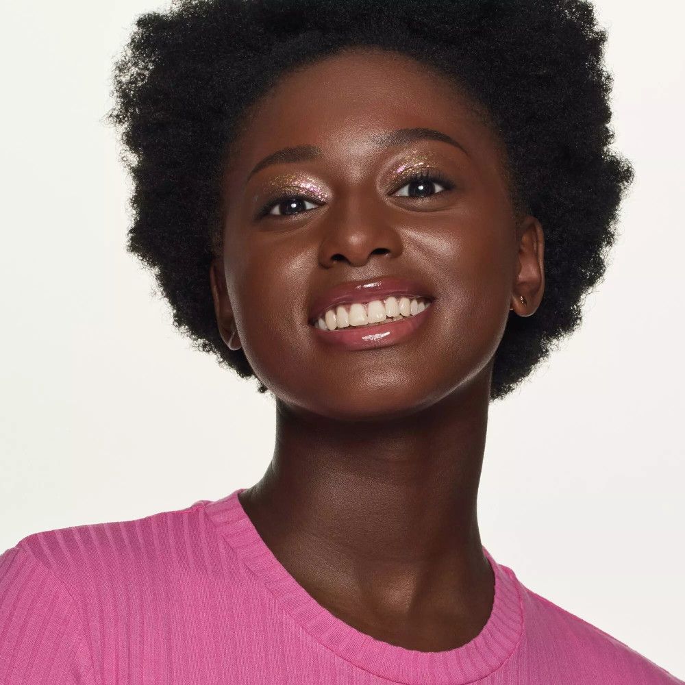 Ritratto di una donna con pelle abbronzata e labbra rosa. Indossa un top rosa e sorride.