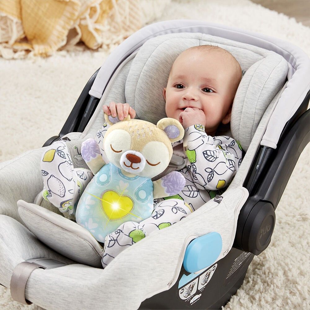 Bambino nel seggiolino auto tiene orsetto con cuore. Orsetto beige, vestito blu. VTech.