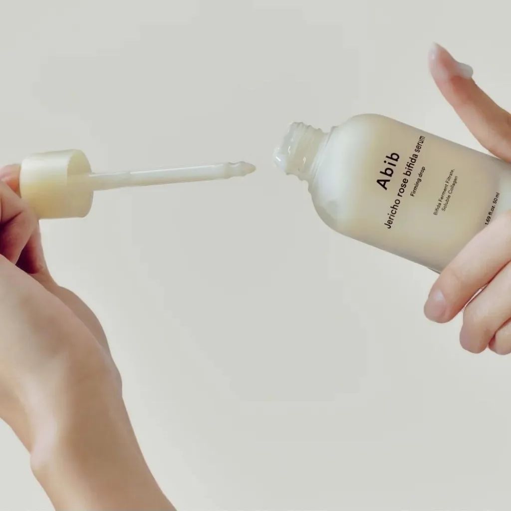 Mani che tengono un flacone di siero e un contagocce. Flacone con scritta: Abib Jericho Rose Bifida Serum Firming Drop. Il contagocce preleva il siero.
