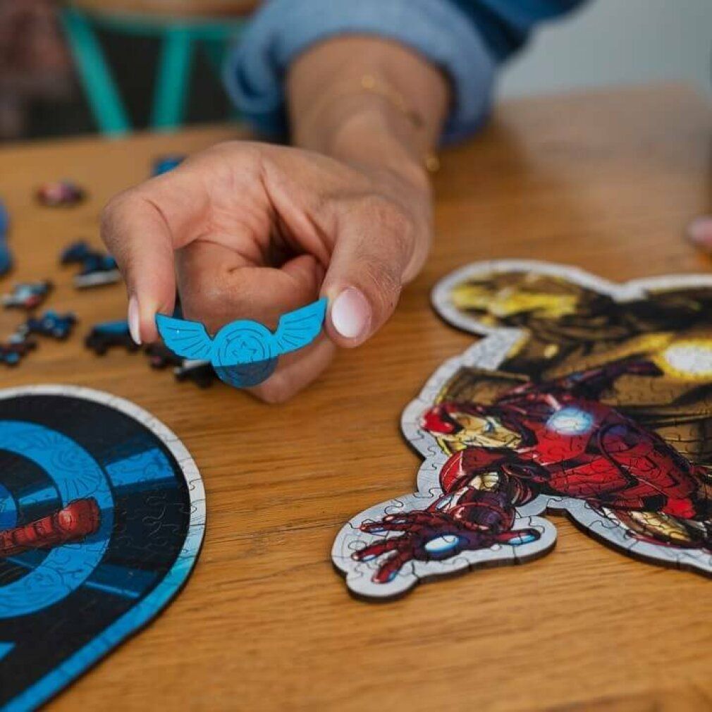 Mano che tiene un pezzo di puzzle blu con motivo ad ali. Accanto, pezzi di puzzle con motivo di Iron Man. Superficie in legno.