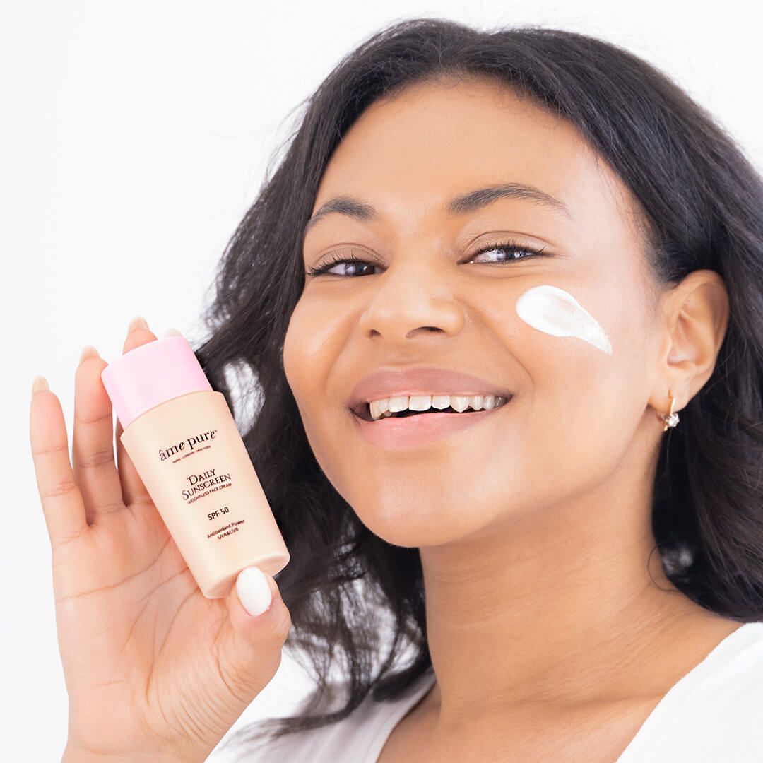 Donna con flacone Âme Pure Daily Sunscreen e crema solare sulla guancia, SPF 50.