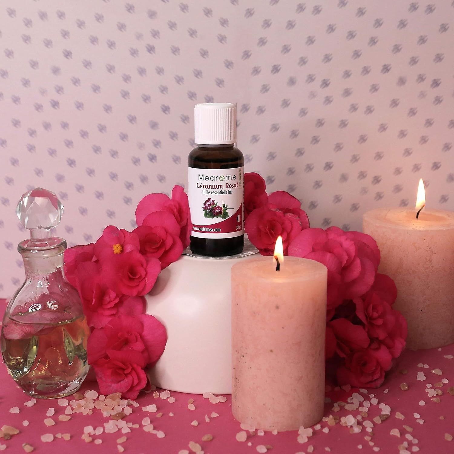 Flacone di vetro marrone con tappo bianco, circondato da fiori rosa e candele. Scritta sull'etichetta.