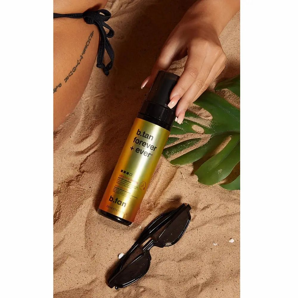 Flacone di mousse autoabbronzante sulla sabbia. Confezione nera e oro. Testo: b.tan forever + ever. Occhiali da sole e foglia accanto. Mano che tiene il flacone.