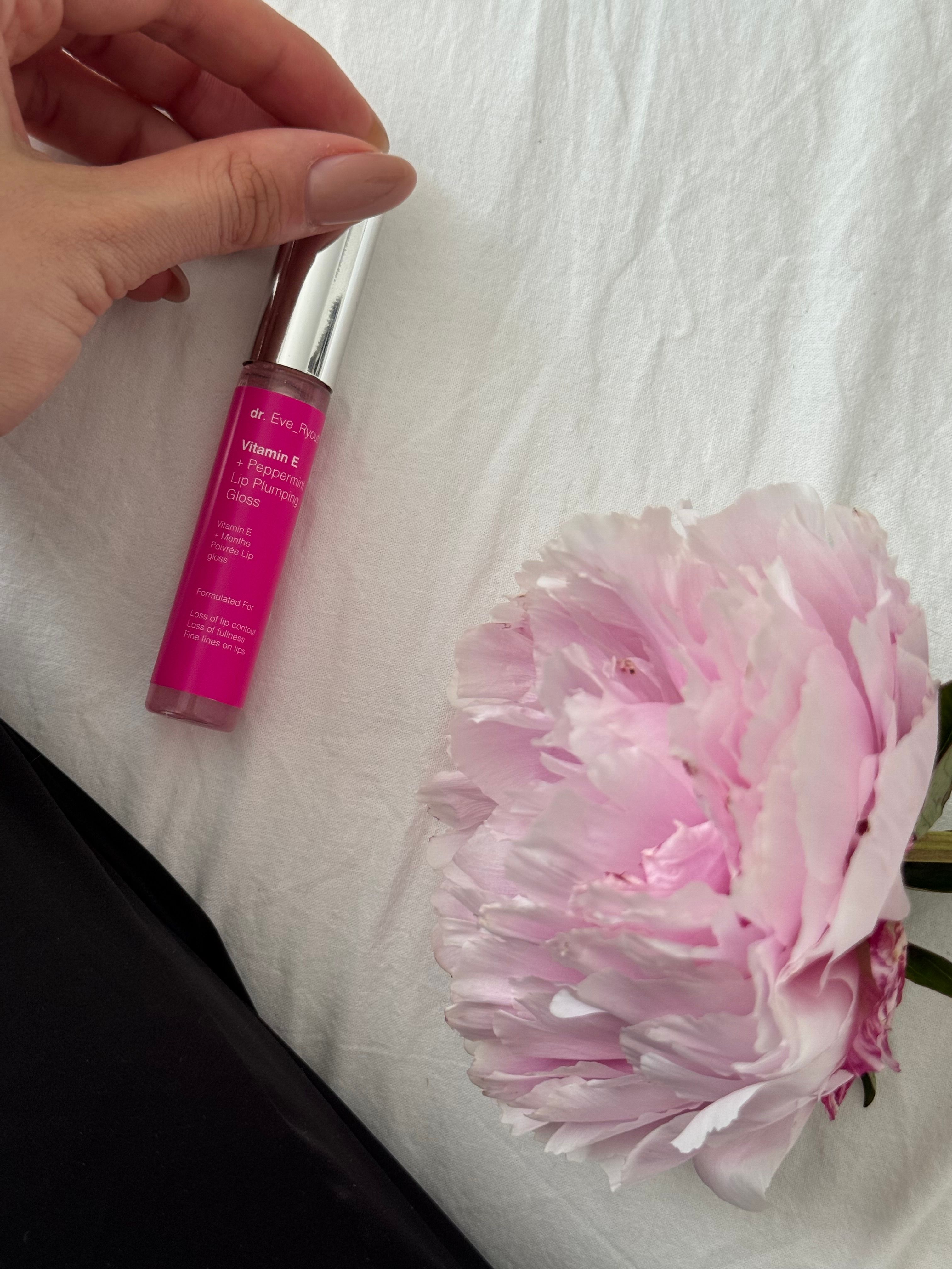 Un tubo di lucidalabbra accanto a un fiore. Liquido rosa in un contenitore trasparente con tappo argentato. Scritta: Dr. Eve_Ryouth, Vitamina E + Menta piperita.