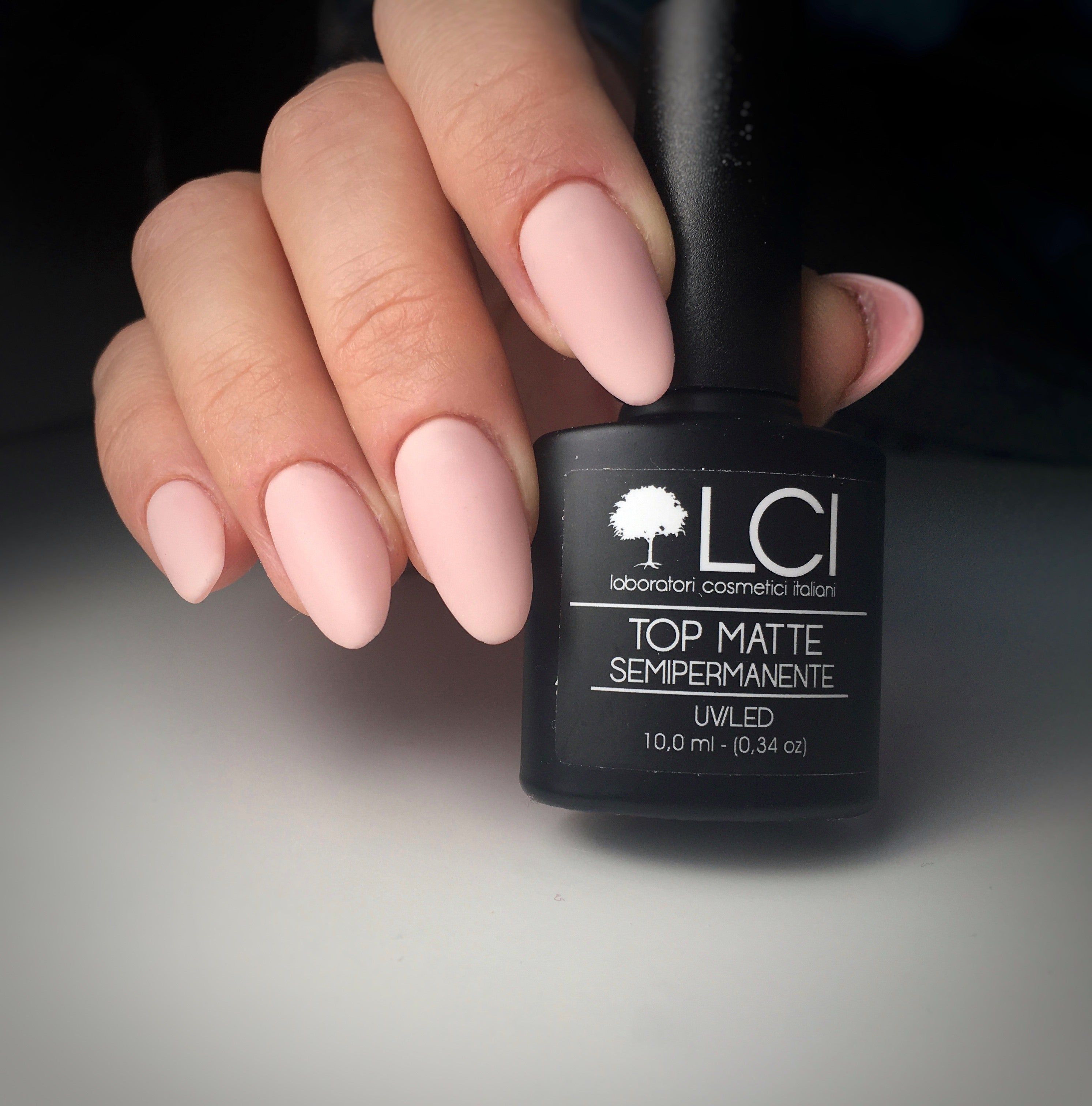 Mano con unghie smaltate e flacone di smalto. Flacone con tappo nero, etichetta con logo e testo. Top Matte.