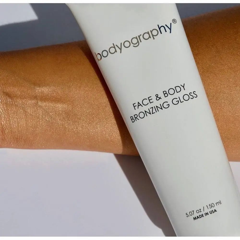 Tubo bianco BODYOGRAPHY FACE & BODY BRONZING GLOSS. Applicazione su braccio abbronzato. 5.07 oz / 150 ml.