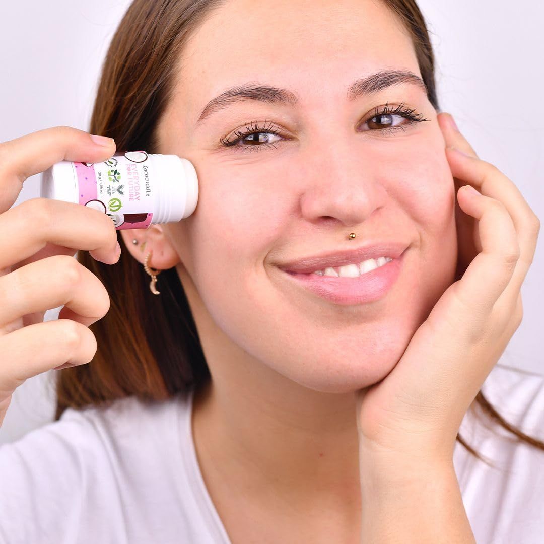 Donna tiene un contenitore cilindrico sulla guancia. Etichetta rosa con scritta: EVERYDAY FOR FUTURE. Applicazione di siero viso.