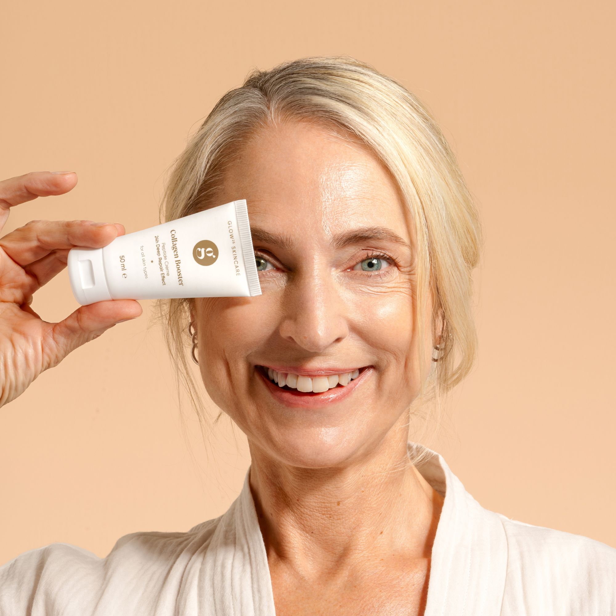 Donna tiene un tubetto di crema davanti all'occhio. Sul tubetto: Collagen Booster, 50 ml.