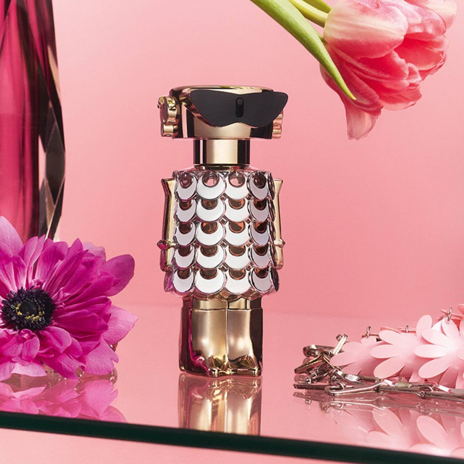 Flacone dorato a forma di robot con elementi argentati. Su superficie rosa con fiori. Occhiali da sole neri sulla testa.