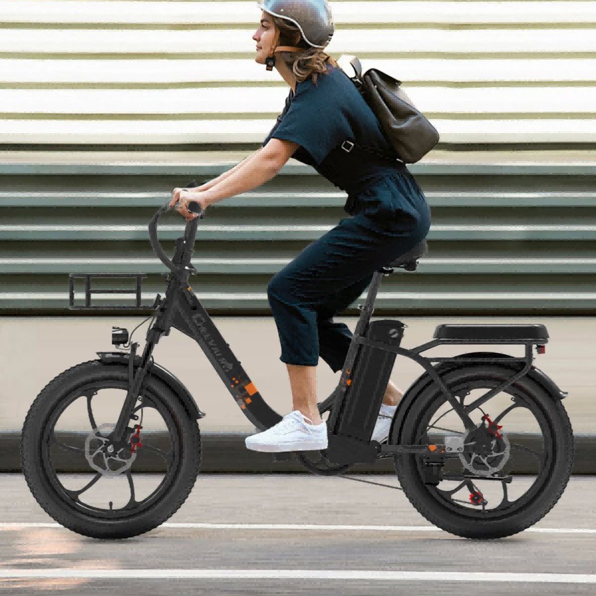 Persona in bicicletta elettrica nera. Indossa casco e zaino. Sfondo: edificio a strisce orizzontali.