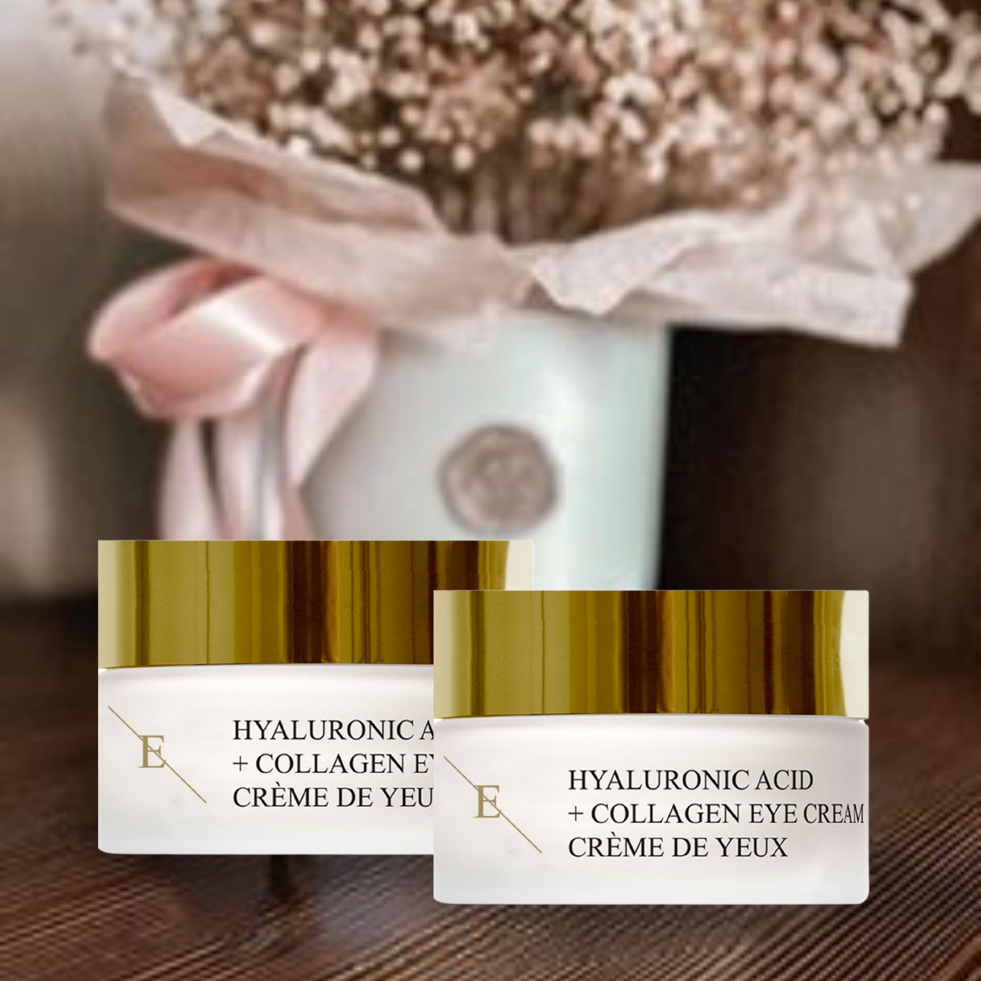 Due vasetti di crema per occhi con coperchi dorati. Scritta: Acido Ialuronico + Collagene Eye Cream. Crème de Yeux. Logo 'E'. Su un tavolo, con fiori sullo sfondo.