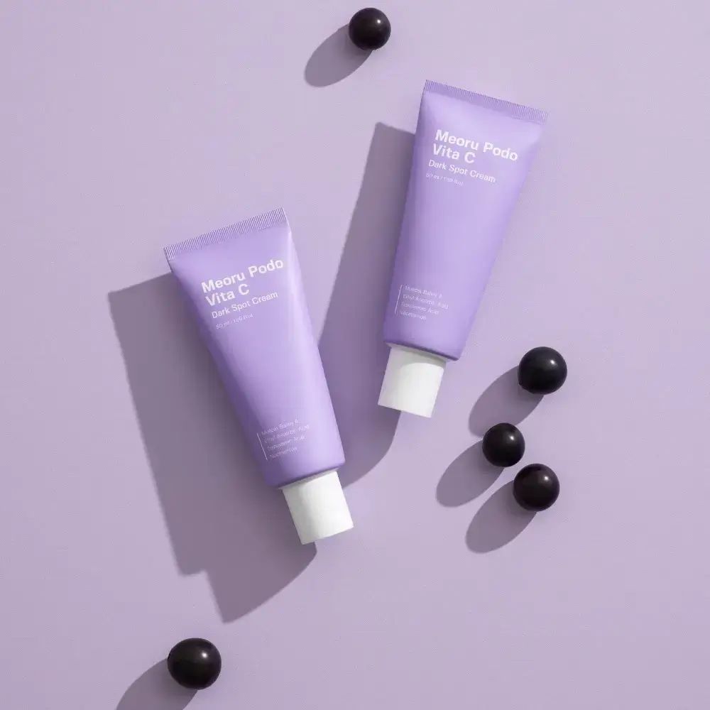 Due tubi viola 'Meoru Podo Vita C Dark Spot Cream' e uva nera su sfondo viola.
