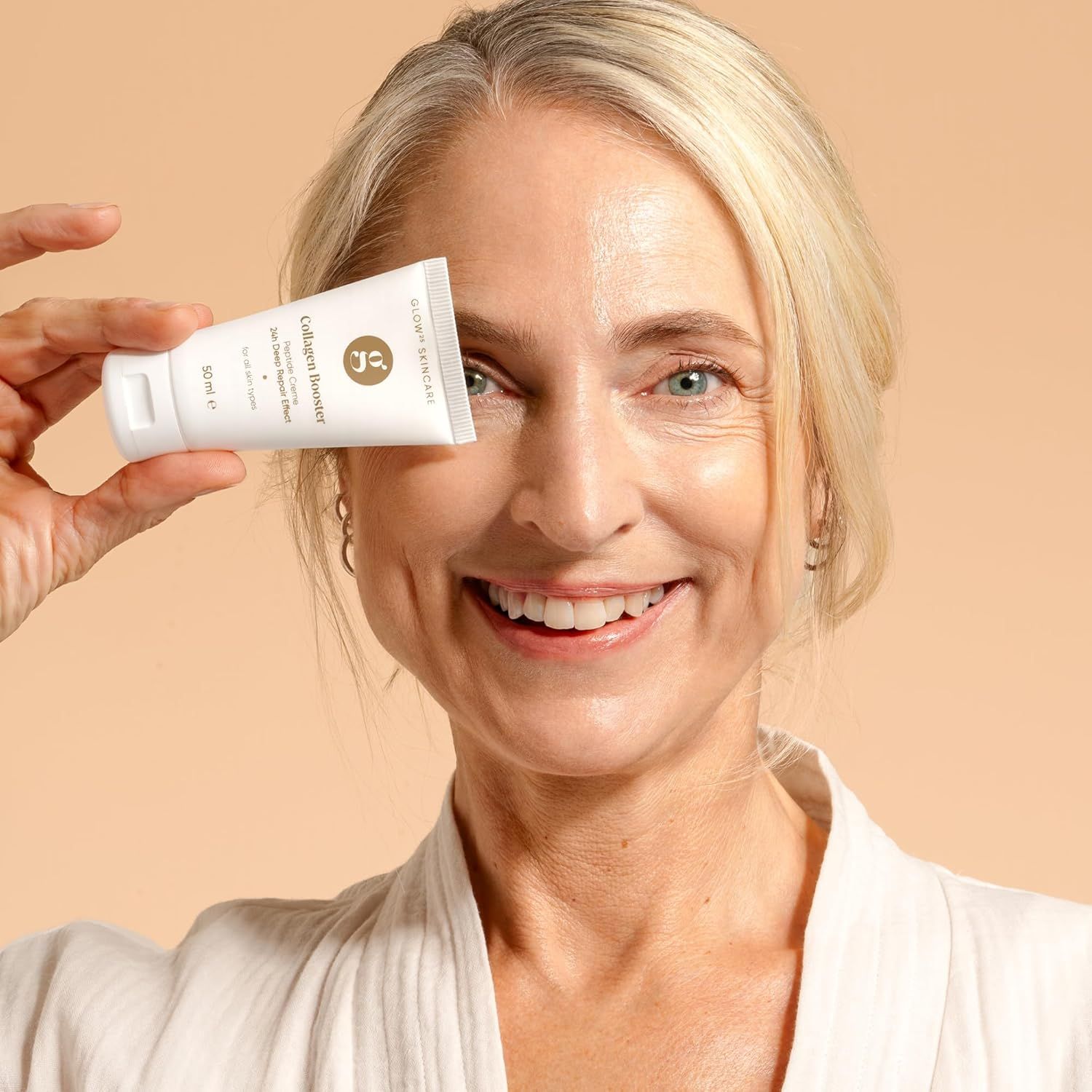 Una donna tiene un tubo bianco "Glow Skincare Collagen Booster" davanti all'occhio. Sul tubo si legge "50 ml". La donna sorride. Indossa un accappatoio bianco.