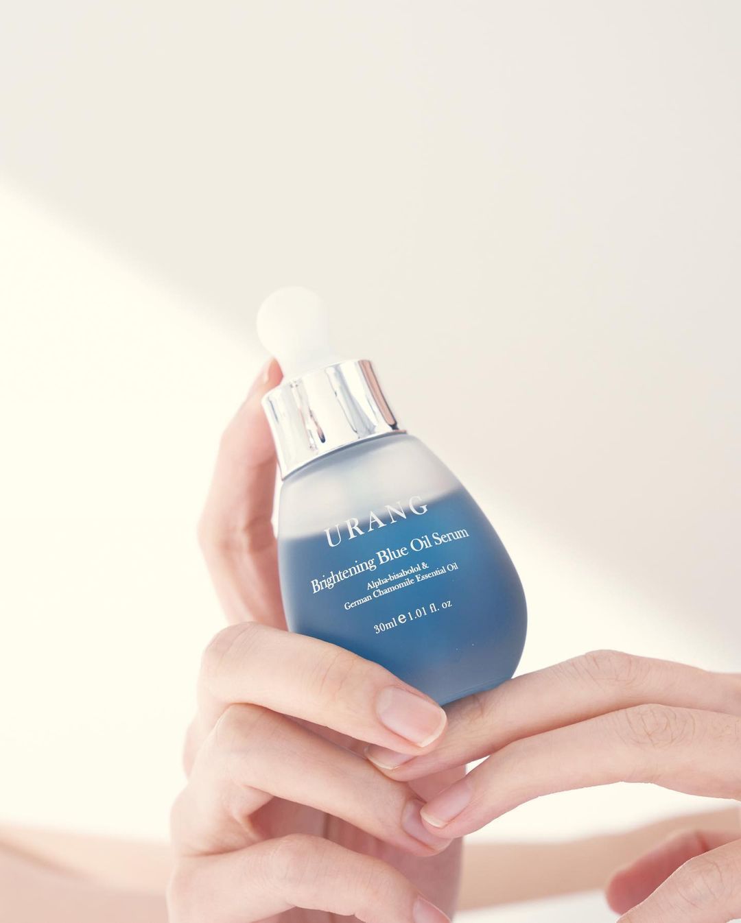 Mani che tengono flacone di siero. Scritta: URANG Brightening Blue Oil Serum. Pipetta visibile.