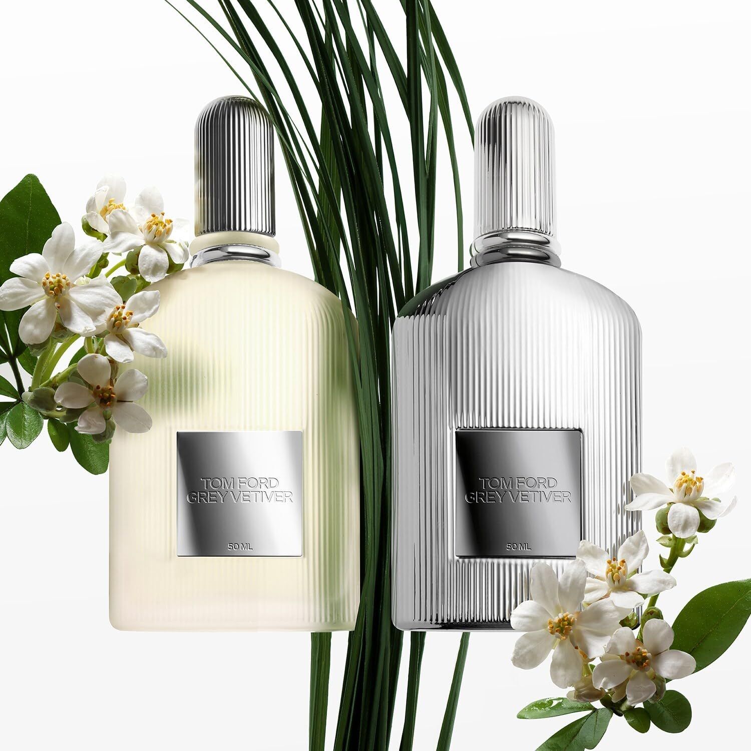 Due flaconi grigi con superficie a coste e tappo argentato. Etichette rettangolari con testo: TOM FORD GREY VETIVER. Con fiori.