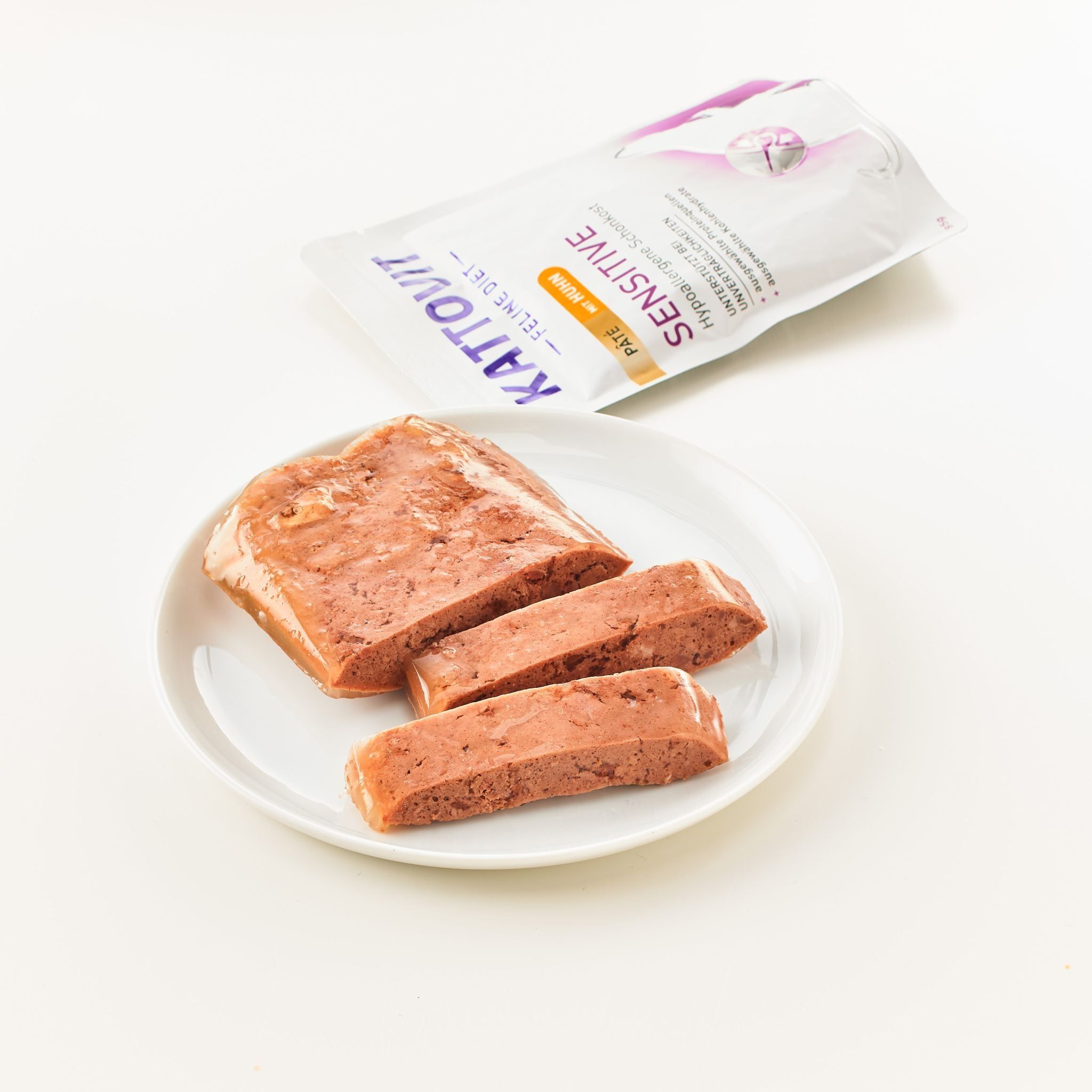 Kattovit Sensitive Paté Huhn su piatto. Paté a forma, tagliato in tre pezzi. Busta sullo sfondo.