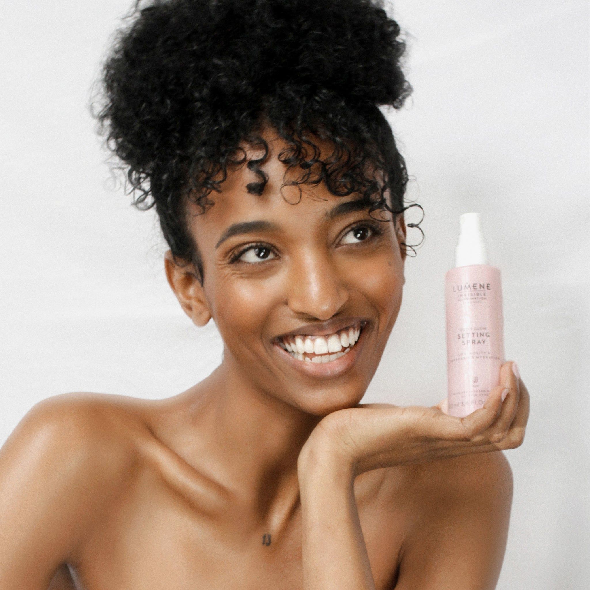 Donna tiene flacone spray rosa. Scritta: Lumene INVISIBLE ILLUMINATION Dewy Glow Setting Spray. Donna sorride. Sfondo: bianco.