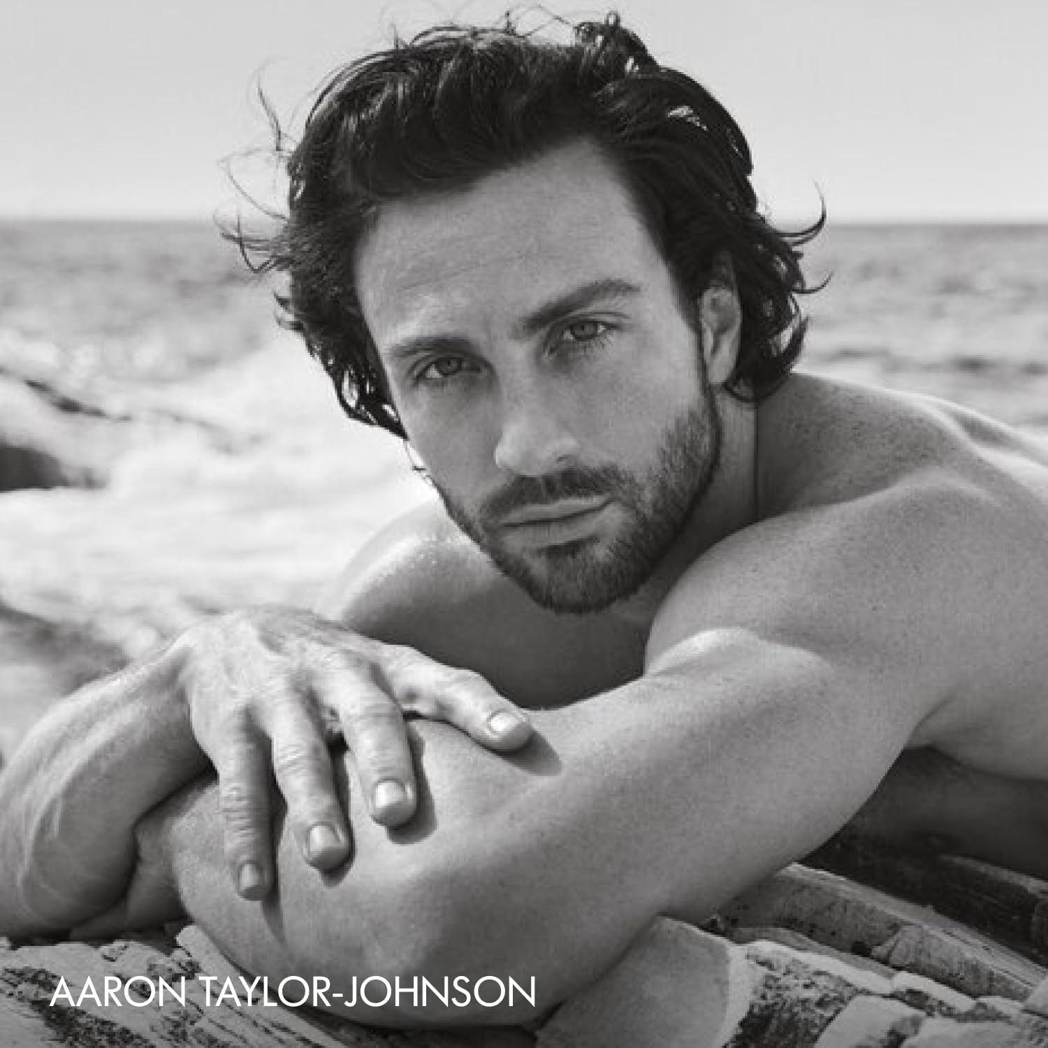 Ritratto in bianco e nero di un uomo sulla spiaggia. L'uomo è sdraiato sulle rocce e guarda la fotocamera. Testo: Aaron Taylor-Johnson.