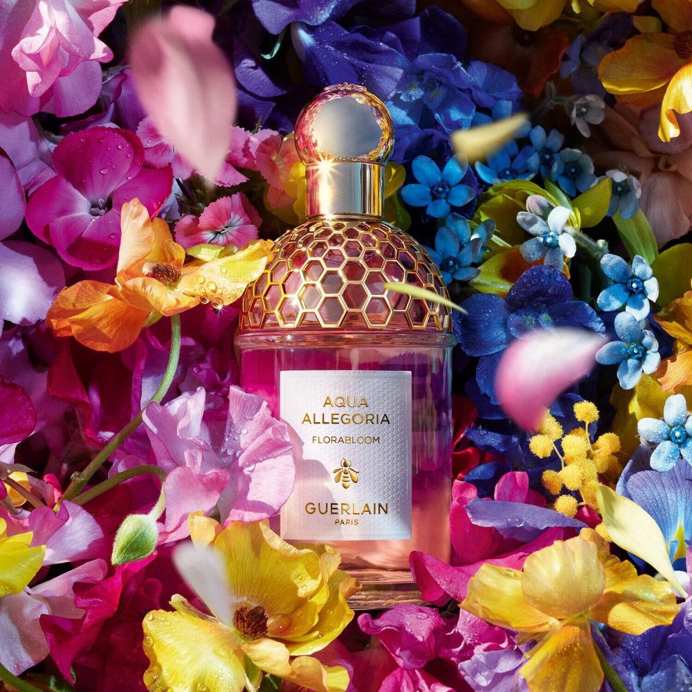Flacone di profumo con tappo dorato, circondato da fiori colorati. Scritta: Aqua Allegoria Florabloom, GUERLAIN.