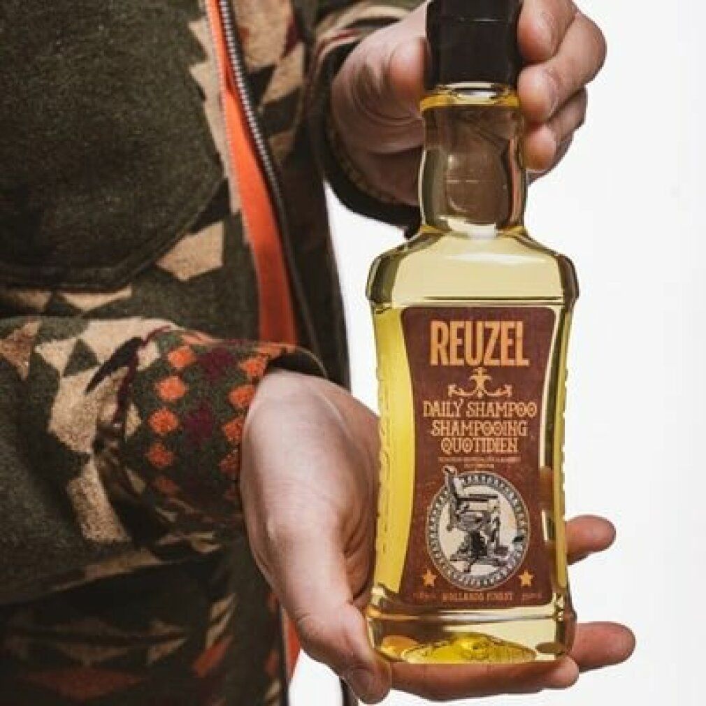 Mano che tiene un flacone di Reuzel Daily Shampoo. Etichetta marrone, tappo nero. Liquido giallo visibile.