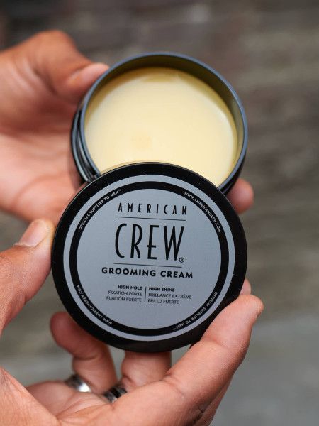 Barattolo aperto "AMERICAN CREW GROOMING CREAM" tenuto in mano. Crema visibile. Testo: "HIGH HOLD, HIGH SHINE".