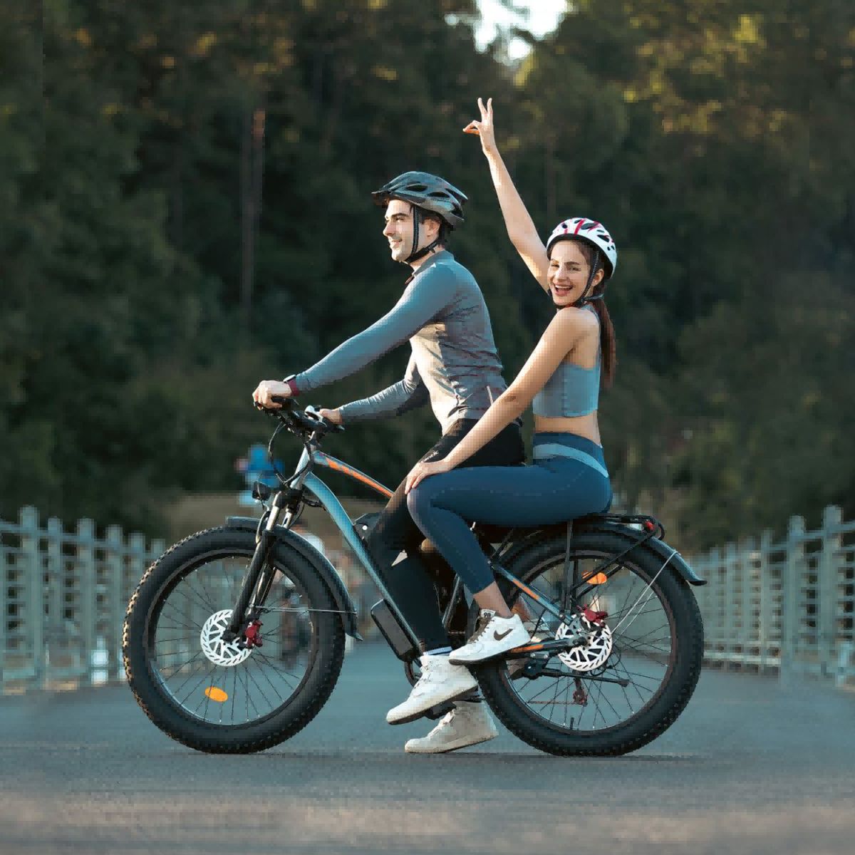 Due persone in sella a un'e-bike fat bike grigia. Indossano caschi. Sfondo: alberi e ponte.