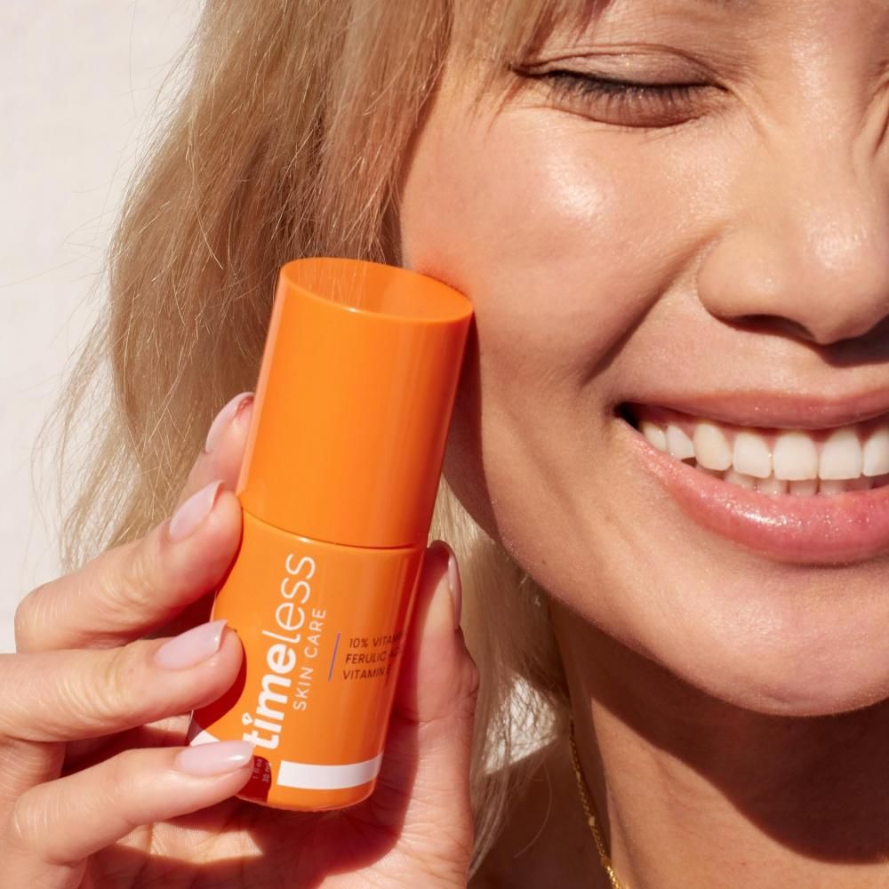 Donna tiene in mano un flacone arancione. Scritta: TIMELESS SKIN CARE, 10% Vitamina C, Acido Ferulico, Vitamina E. La donna sorride.