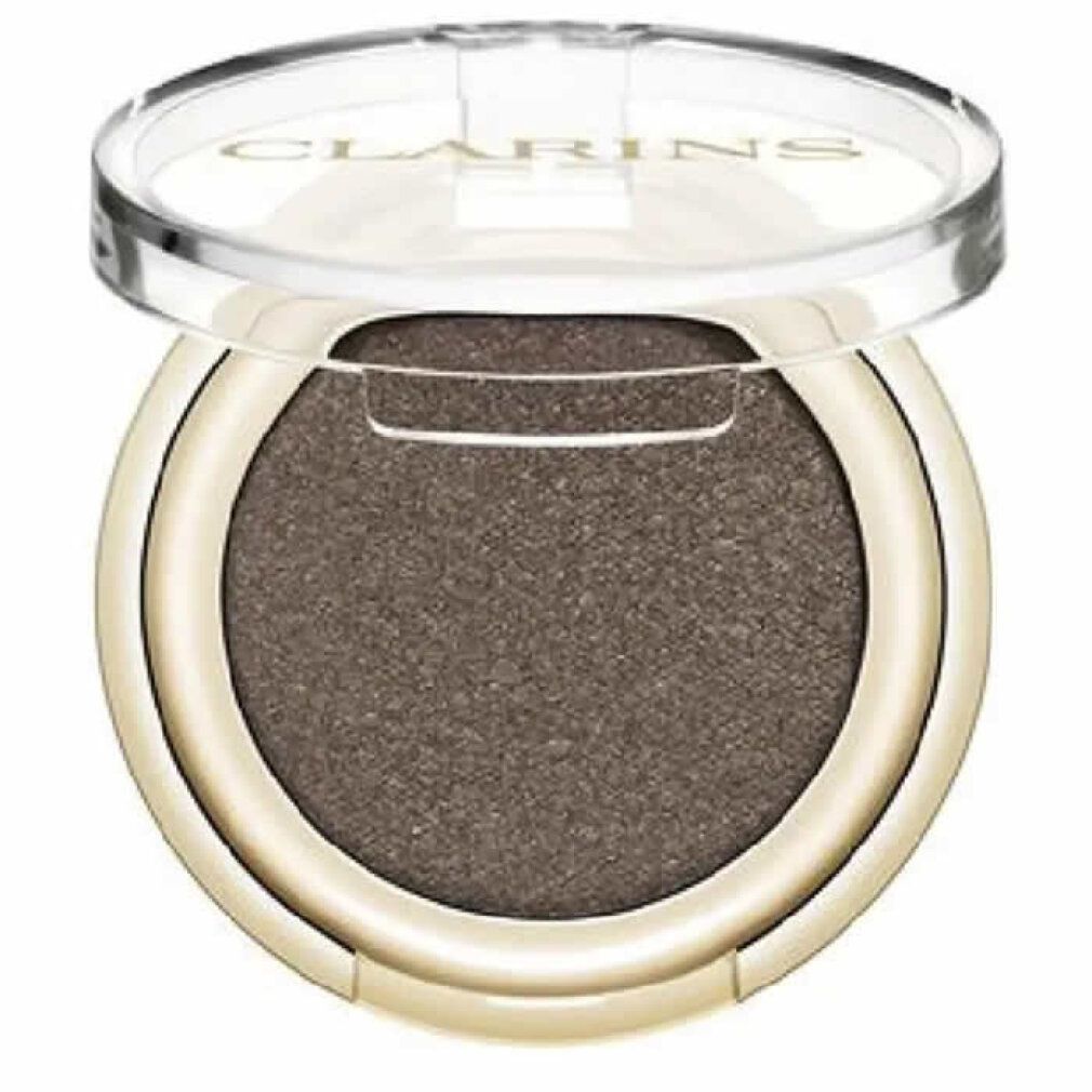 Contenitore rotondo dorato per ombretto con coperchio trasparente. L'ombretto è marrone. Il marchio Clarins è visibile sul coperchio.