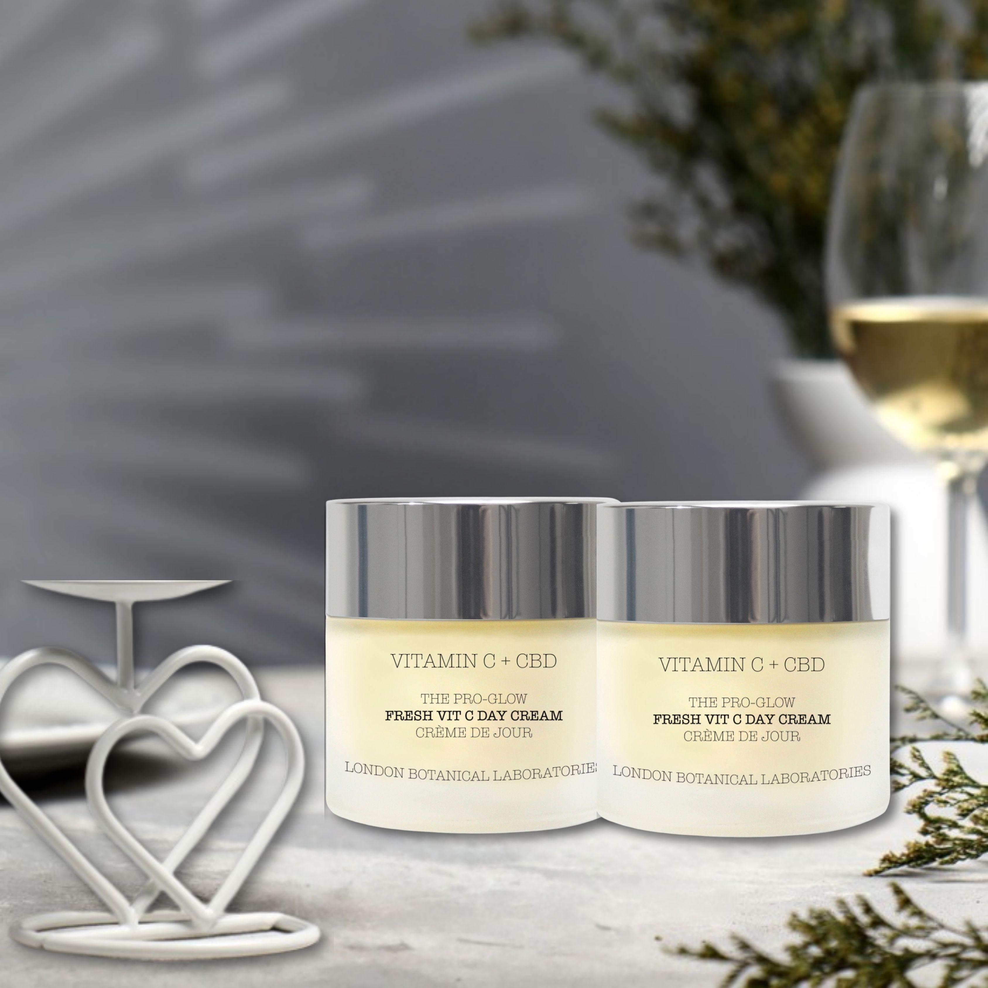 Due vasetti di crema con coperchi argentati. Scritta: Vitamin C + CBD, The Pro-Glow Fresh Vitamin C Day Cream. Su sfondo chiaro.
