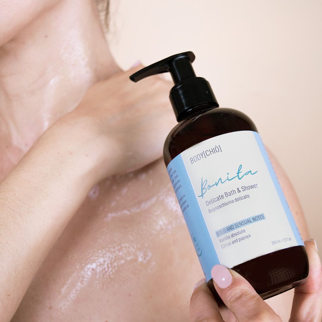 Flacone marrone con dosatore nero. Scritta: BODYCHIÒ, Bonita, Delicate Bath & Shower. Persona applica il prodotto.