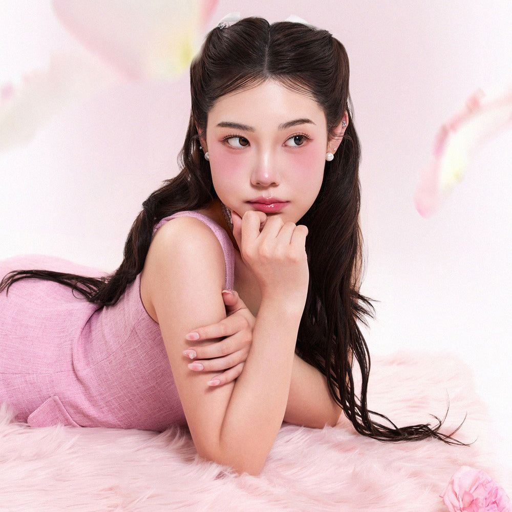 Una donna sdraiata su un tappeto rosa, indossa abiti rosa e ha il fard sulle guance. Guarda a destra.