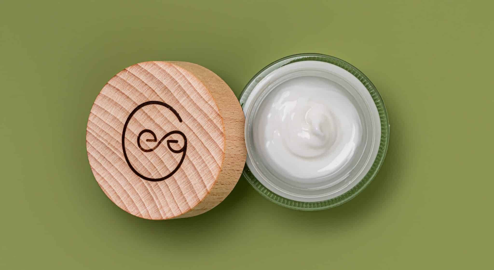 Vista dall'alto di vasetto di crema e coperchio in legno. Coperchio con logo. Crema nel vetro. Sfondo verde.