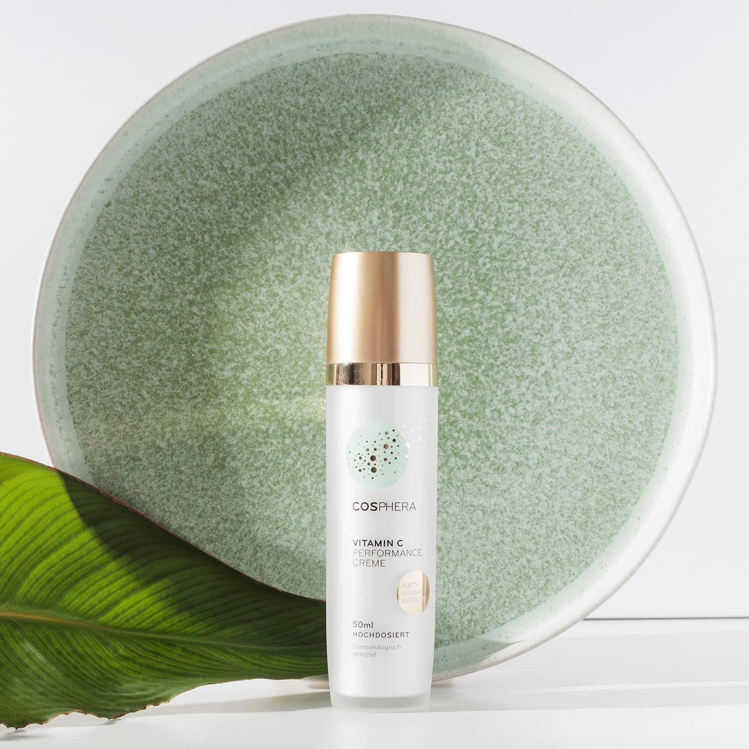 Flacone di crema Cosphera Vitamina C accanto a un piatto verde e una foglia. Dispenser a pompa con tappo dorato.
