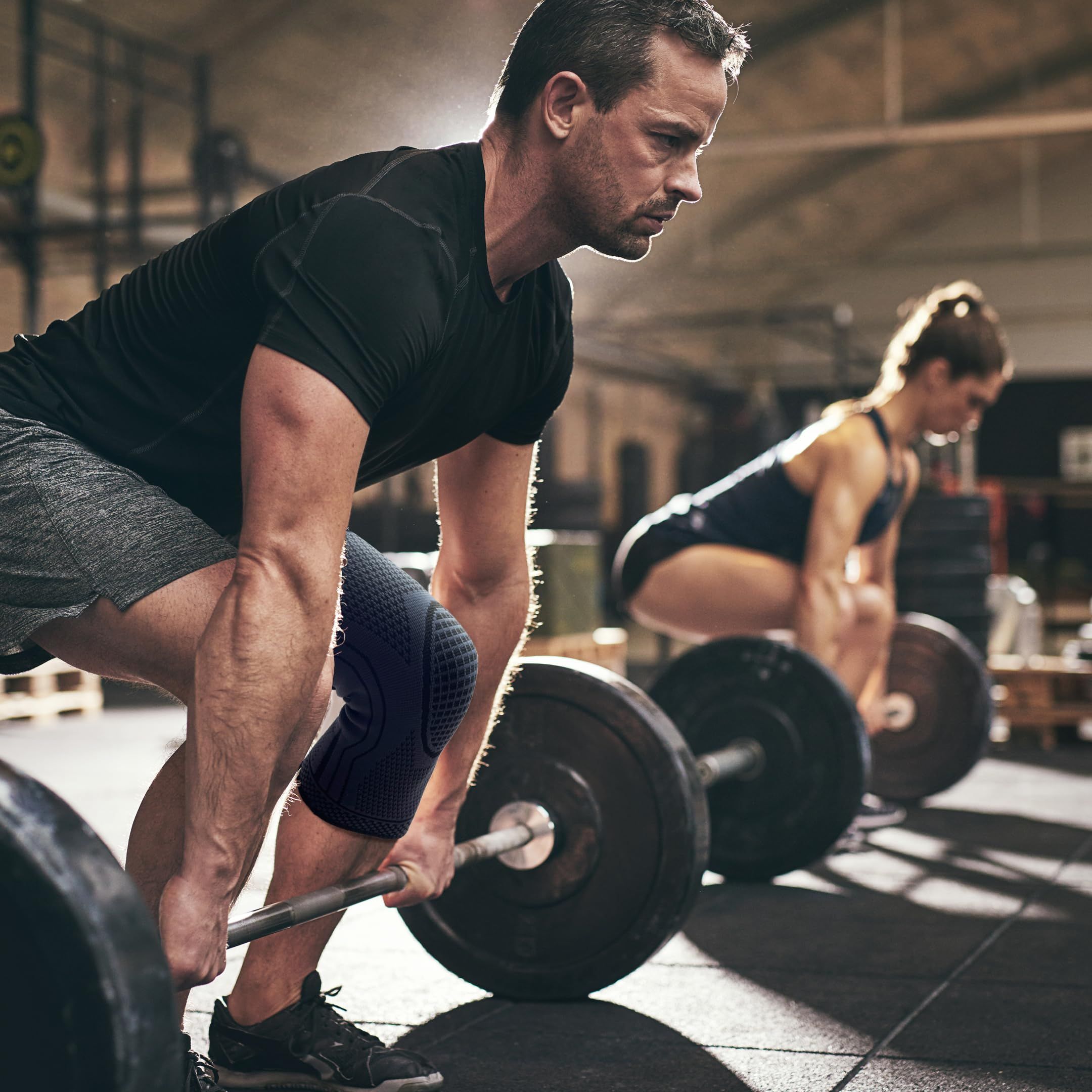 Due persone sollevano pesi. Entrambi indossano ginocchiere. Ambiente palestra.